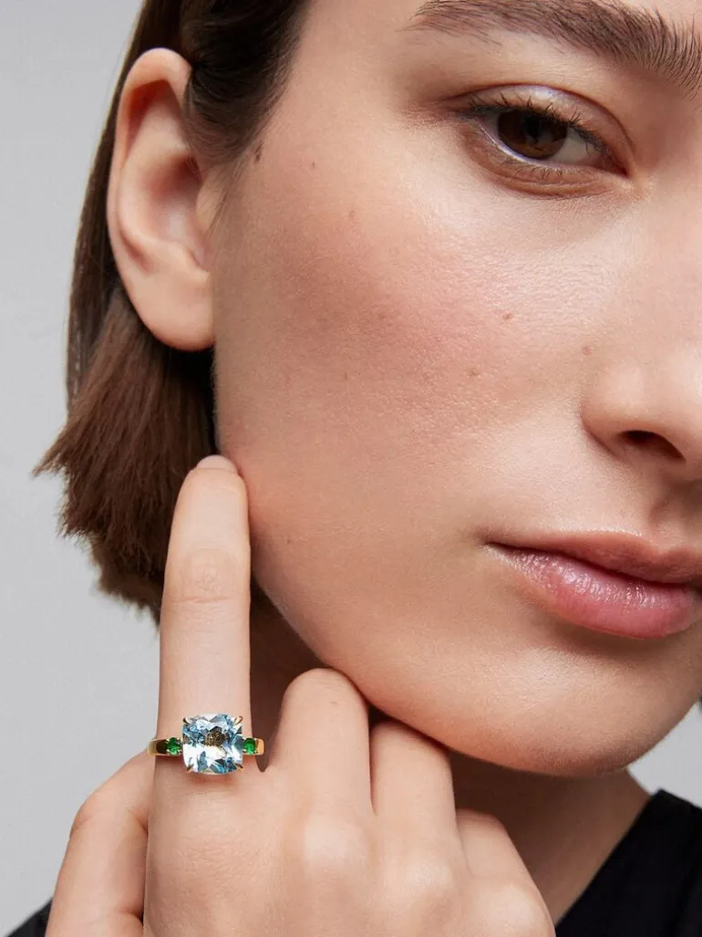 Anillo tresillo de oro amarillo de 18K con topacio azul sky en talla cushion de 4,88 cts y 2 esmeraldas verdes en talla brillante